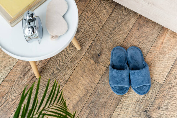 вид сверху на мягкие тапочки возле прикроватного столика с винтажными будильниками, маской для сна и книгами