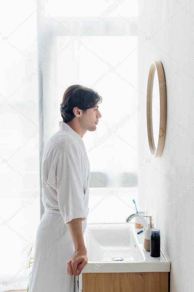 side view of man in bathrobe looking in mirror near toiletries on sink