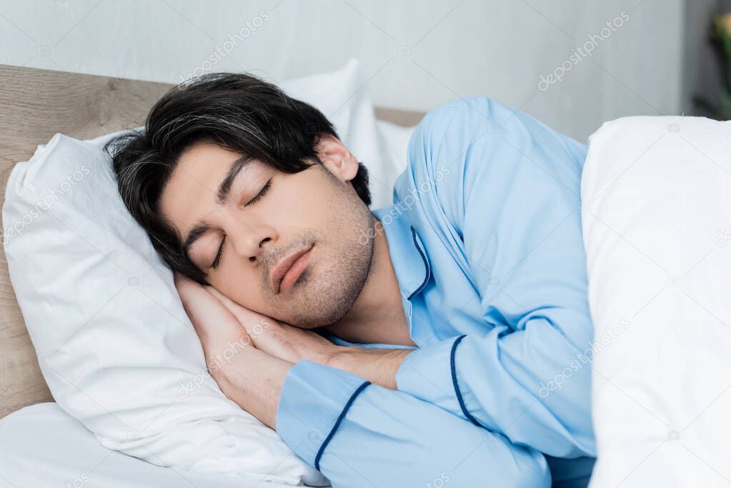 young brunette man in blue pajamas sleeping on white bedding