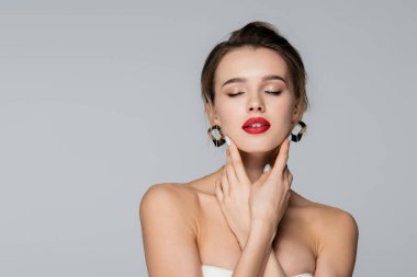 Çıplak omuzlu genç bir kadın yüzüne dokunurken gözleri kapalı gri renkte poz veriyor.