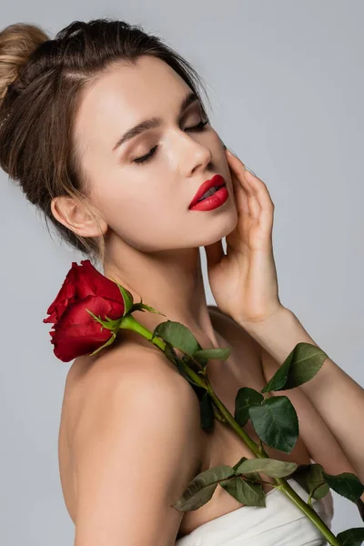 Urocza Kobieta Czerwoną Różą Dotykająca Twarzy Podczas Pozowania Zamkniętymi Oczami — Zdjęcie stockowe