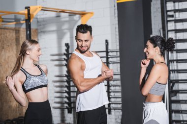 Gülümseyen sporcu spor salonunda ırklar arası kadın sporcuların yanında kaslarını gösteriyor. 