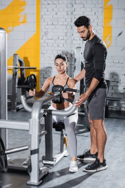 Orta Doğu sporcusunun yanında duran Arap antrenör spor salonunda karın kası çatırdatma makinesi eğitimi alıyor. 