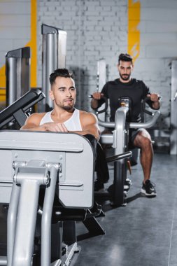 Spor salonundaki bulanık Arap arkadaşının yanında bacak pres makinesinde gülümseyen sporcu eğitimi 