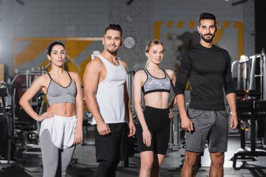 Spor giyim kuşağındaki neşeli ırklar arası insanlar spor salonundaki kameraya bakıyor. 