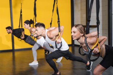 Blonde sportswoman training with suspension straps near blurred interracial friend in gym  clipart