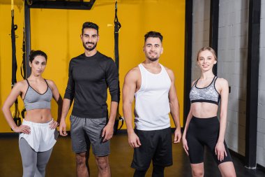 Spor kıyafetlerinde gülümseyen, spor salonunda kameraya bakan çok ırklı arkadaşlar. 