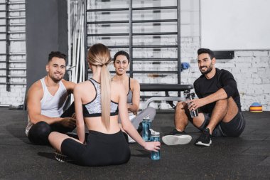 Spor şişeleriyle gülümseyen çok ırklı genç insanlar spor salonunda arkadaşlarıyla konuşuyor. 