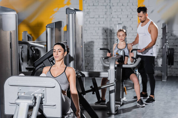 Middle east sportswoman training on leg press machine near blurred friends in gym 