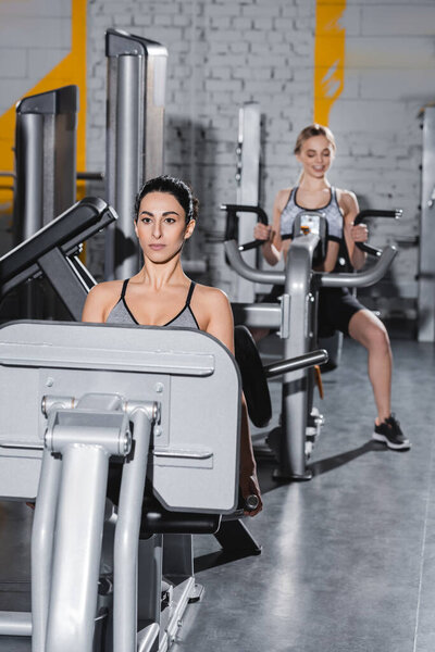 Young middle east sportswoman working out on leg press machine near blurred friend in gym 