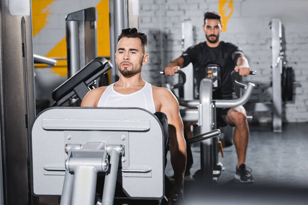 Sportsman training on leg press machine near blurred muslim friend in gym 