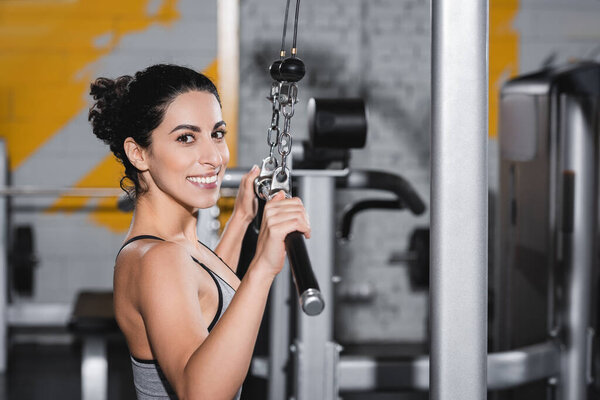 Cheerful middle east sportswoman looking at camera near lat pulldown machine in gym 