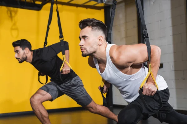 Athletic sportsman training with suspension straps near muslim man in gym