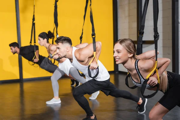 Blonde sportswoman training with suspension straps near blurred interracial friend in gym
