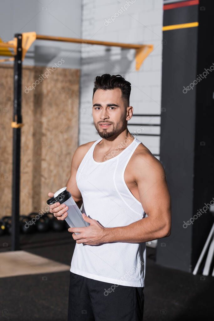Smiling sportsman holding sports bottle and looking at camera in gym