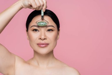 Asian woman with naked shoulders massaging forehead with jade roller isolated on pink  clipart