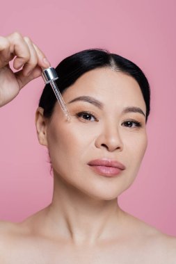 Asyalı kadın pembeye izole edilmiş yüzüne serum sürüyor. 
