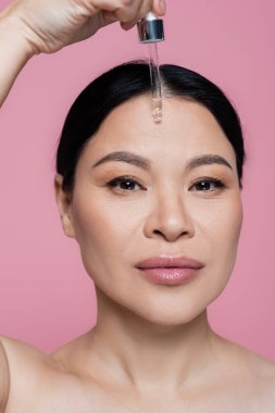 Çıplak omuzlu Asyalı kadın pembeye izole edilmiş yüzüne serum uyguluyor. 