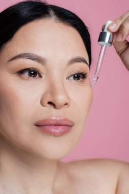 Asyalı kadının pembeye izole edilmiş yüzüne serum uyguladığı yakın plan. 