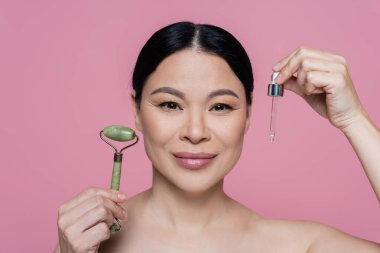 Çıplak omuzlu Asyalı bir kadın, yeşil bigudi ve pembeye izole edilmiş serum tutuyor. 