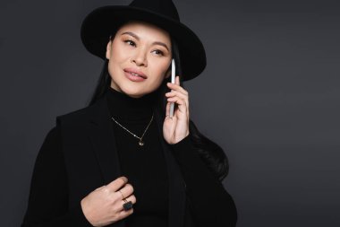 Fötr şapkalı gülümseyen Asyalı kadın cep telefonuyla konuşuyor koyu gri renkte.