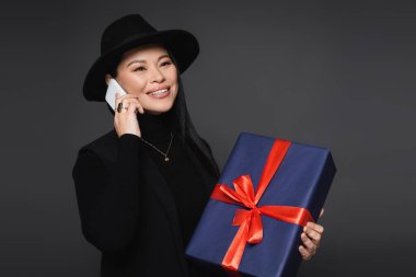 Fötr şapkalı pozitif Asyalı kadın akıllı telefonla konuşuyor ve elinde koyu gri bir hediye tutuyor.