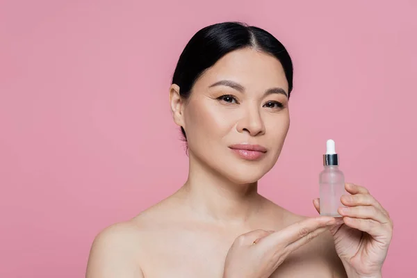 Asian woman with naked shoulders holding serum isolated on pink