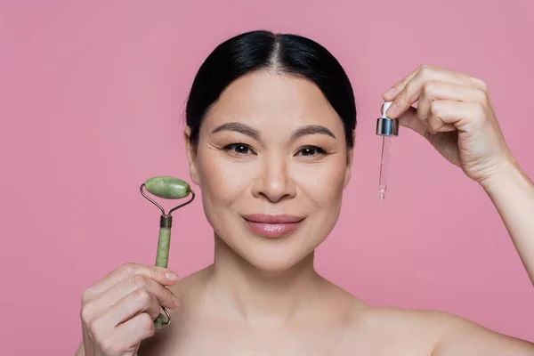 Asian woman with naked shoulders holding jade roller and dropper of serum isolated on pink