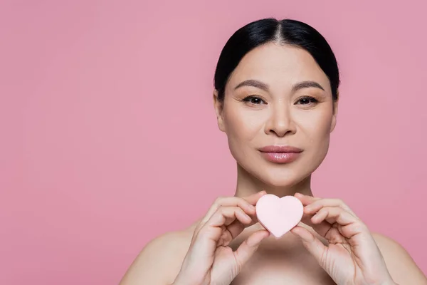 Asian woman with naked shoulders holding heart shaped beauty blender isolated on pink