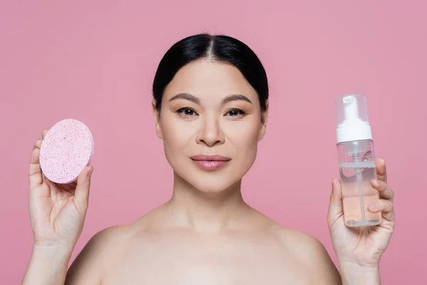 Asian woman with naked shoulders holding sponge and cleansing foam isolated on pink