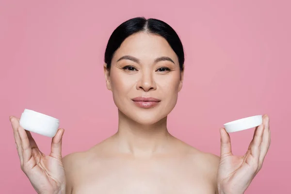 Asian woman with naked shoulders holding container with cosmetic cream isolated on pink