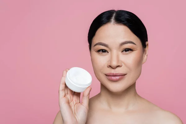 Asian woman with naked shoulders holding cream isolated on pink