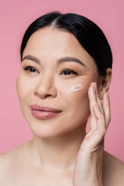 Asian woman applying cream on cheek isolated on pink