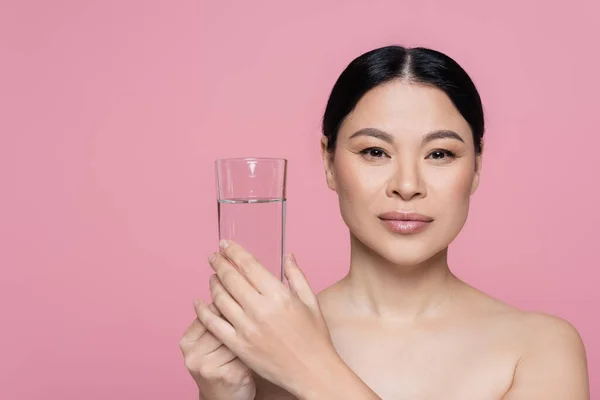 Asian woman with naked shoulders holding glass of water isolated on pink