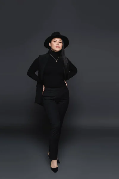 フル長いですアジアの女性でFedora帽子保持手オンヒップ上のダークグレーの背景 — ストック写真