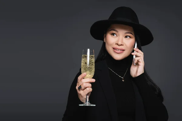 Elegant asian woman in fedora hat talking on smartphone and holding champagne isolated on dark grey