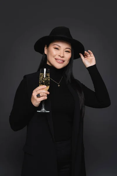 笑容满面的亚洲女人头戴菲洛拉帽 身穿西装 手里拿着一杯深灰色的香槟酒 — 图库照片
