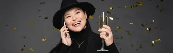 幸せなアジアの女性でフェドーラ帽子話してスマートフォンと開催シャンパン下のConfetti上のダークグレーの背景 バナー — ストック写真