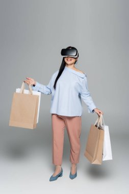 Gri arka planda alışveriş torbaları taşıyan, vr kulaklıklı gülümseyen kadın. 