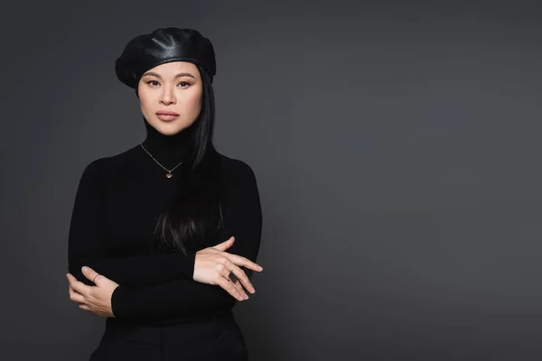Trendy asian model in beret and necklace looking at camera isolated on dark grey