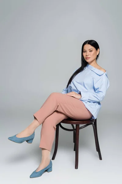 Stylish asian woman positing on chair and looking at camera on grey background