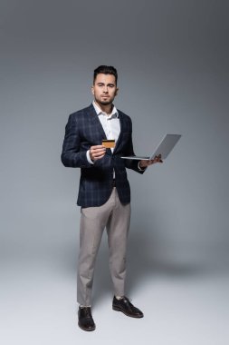Gri üzerinde kredi kartı ve laptop tutan takım elbiseli bir işadamı.