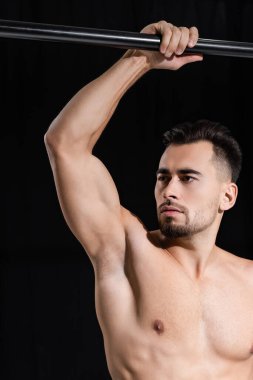 Üstsüz ve kaslı sporcu, siyah üzerine izole edilmiş yatay çubuğun yakınına bakıyor. 