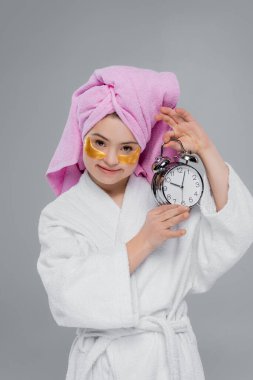 Göz bantlı ve bornozlu kadın alarm saatini gri renkte tutuyor. 