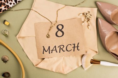 Makyaj fırçasının yanında külot üzerine 8 Mart harfi ve yeşil arka planda topuklu ayakkabı. 