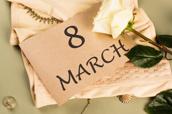 Вид сверху на карточку с 8 Марта на корсете рядом с розой на зеленом фоне 