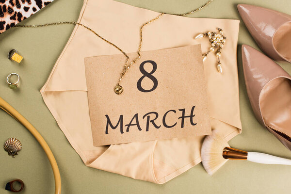 Верхний вид карты с 8 мартовскими надписями на трусиках возле кисти для макияжа и каблуки на зеленом фоне 