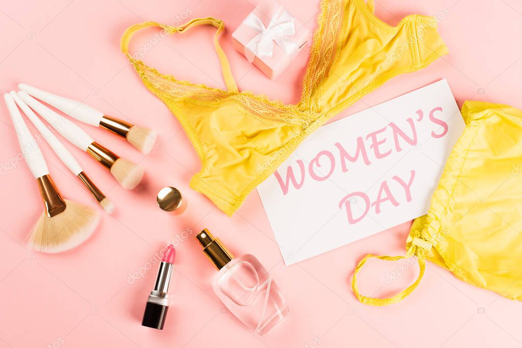 Top view of card with womens day lettering near bra, gift and cosmetic brushes on pink background 