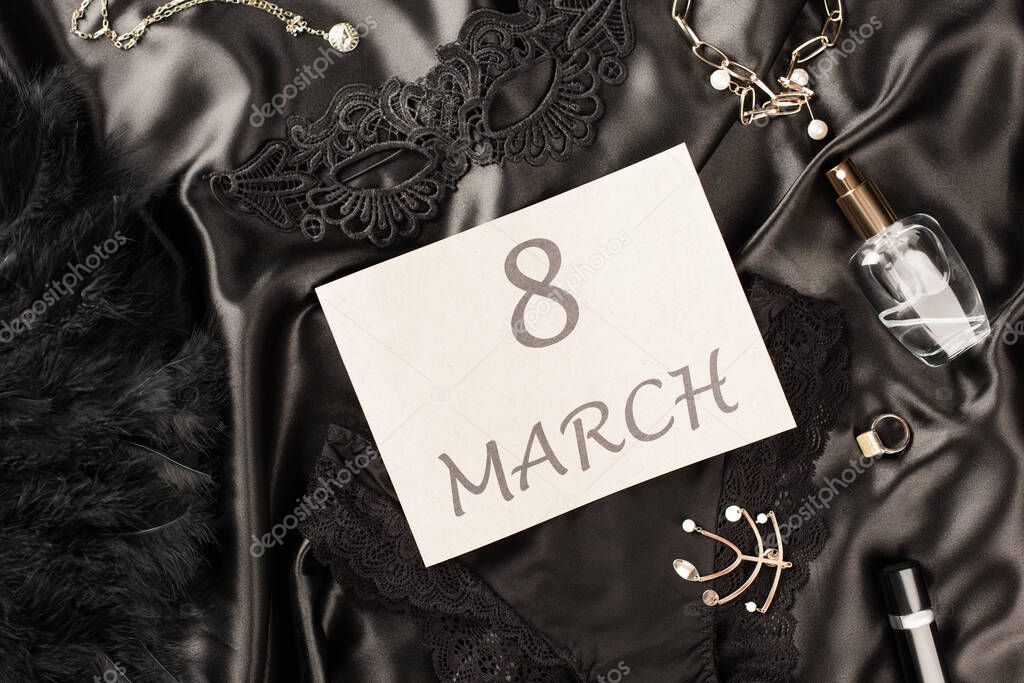 Top view of card with 8 march lettering near panties and sensual mask on black satin background