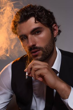Sakallı iş adamı sigara içiyor ve gri arka planda kameraya bakıyor.